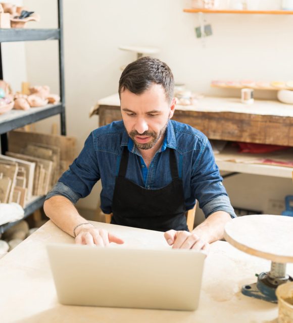 Confident male potter using laptop computer while sitting at table in studio