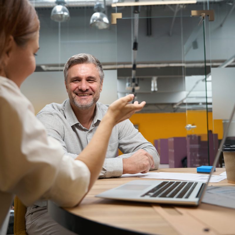 Pretty female sitting at the table near laptop, man in office clothes sitting opposite and talking