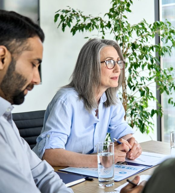 Older business woman leader manager talking to colleagues executive team, diverse partners group at board room meeting. Multicultural professional business people discussing project plan in office.