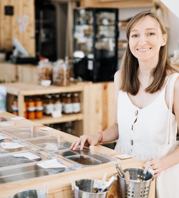 Beautiful girl with cotton bag and glass jars makes conscious shopping in zero waste shop. Woman buying dry goods in plastic free grocery store. Sustainable small businesses. Minimalist lifestyle.